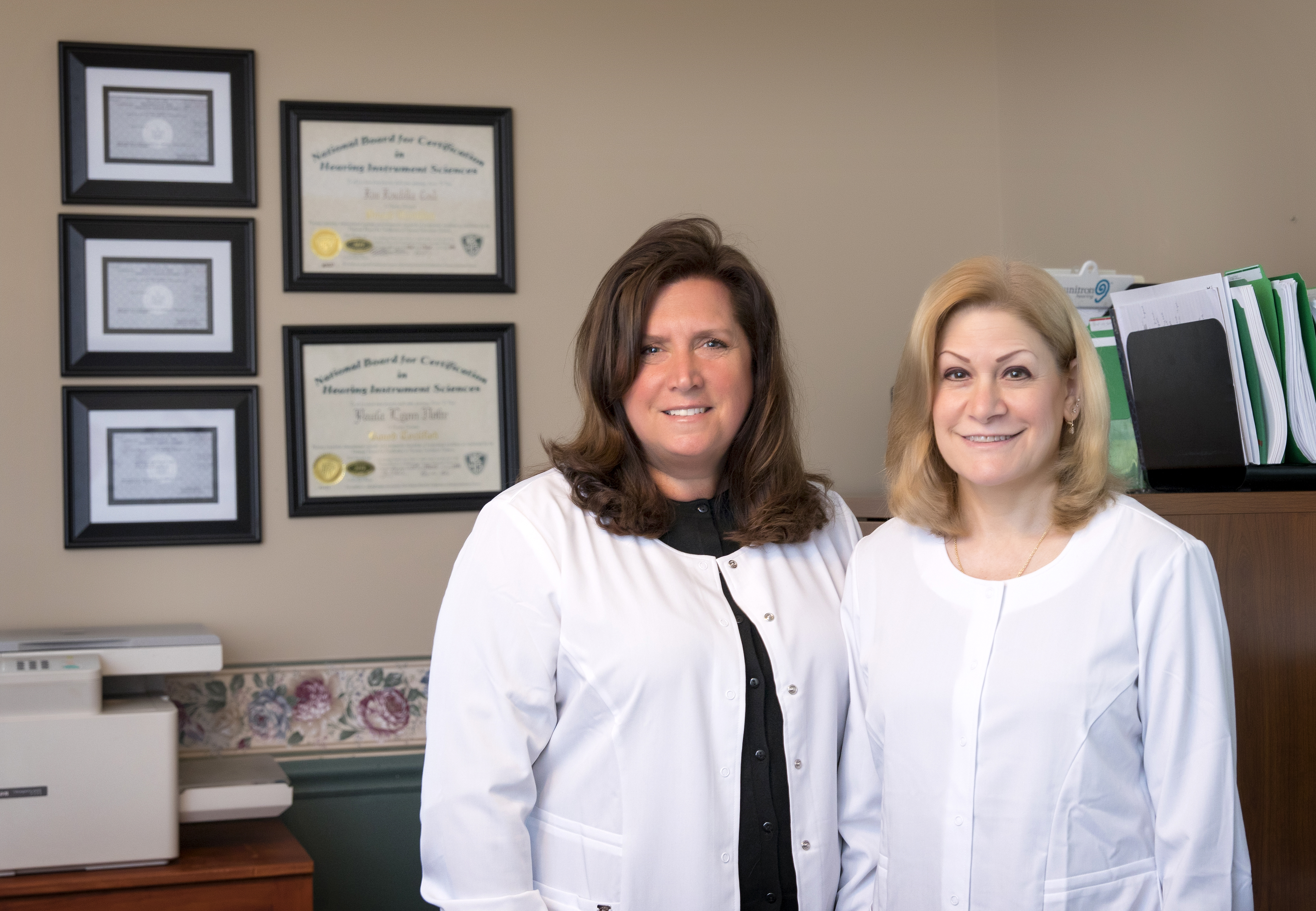 Two women standing in a room with some diplomas on the wall.